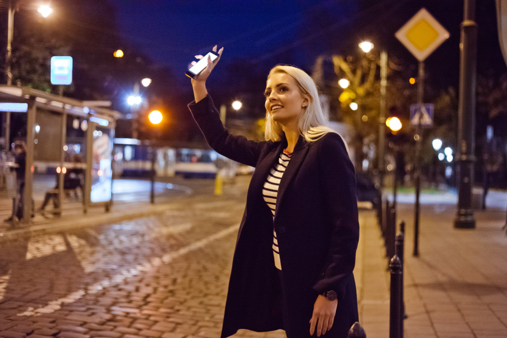 Young blonde woman standing in the city street in the evening, holding smart phone in hand and waiting for taxi or uber.
