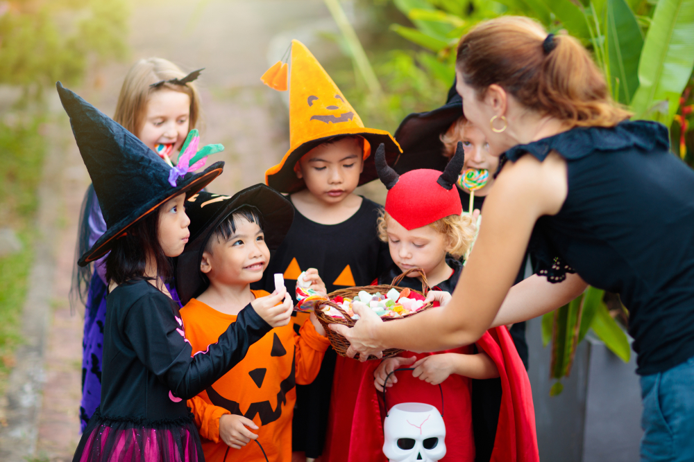 Kids trick or treat on Halloween night. Mixed race Asian and Caucasian children at decorated house door. Boy and girl in witch and vampire costume and hat with candy bucket and pumpkin lantern. 