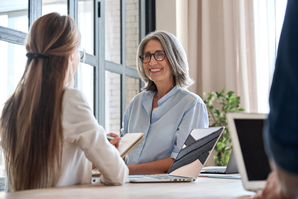 Happy mature middle 60s aged smiling lady boss manager teacher talking to assistant or student meeting in office space. Company team working together with the laptops and assistant writing notes.
