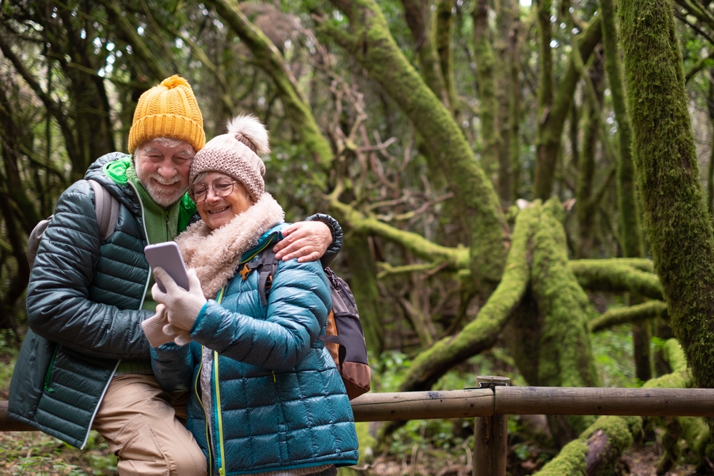 Smiling senior couple enjoying nature outdoors in mountain forest with moss covered trunks. Elderly couple embraced looking at phone. Excursion together in the Garajonay National Park of La Gomera
