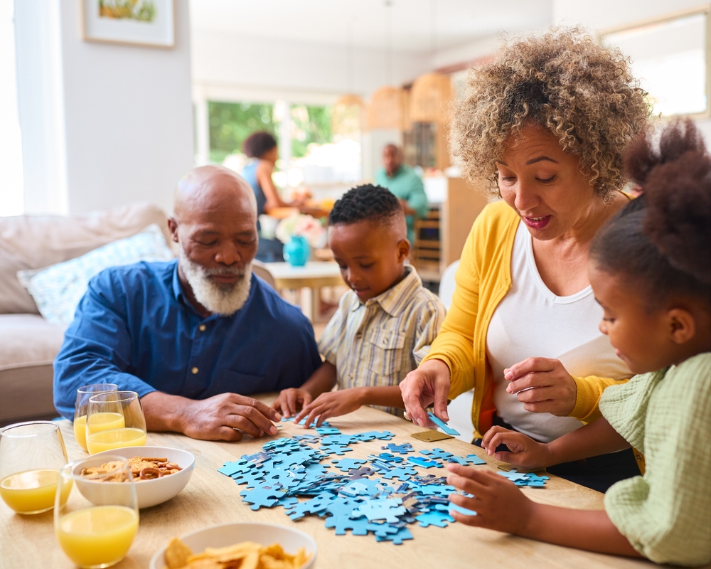 Grandparents With Grandchildren Indoors At Home Doing Jigsaw Puzzle With Parents In Background
