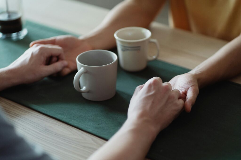 Two people's hands reaching toward each other across a table with coffee cups, showing connection and support.