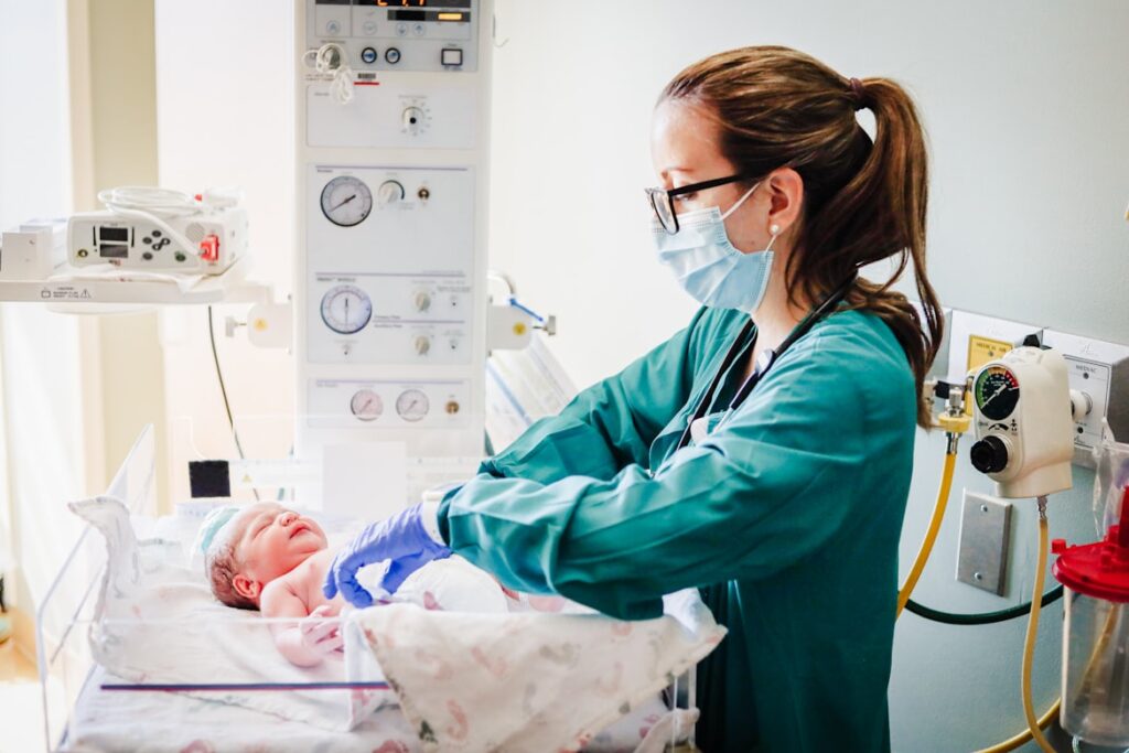 Healthcare worker in green scrubs and mask caring for infant in neonatal intensive care unit.