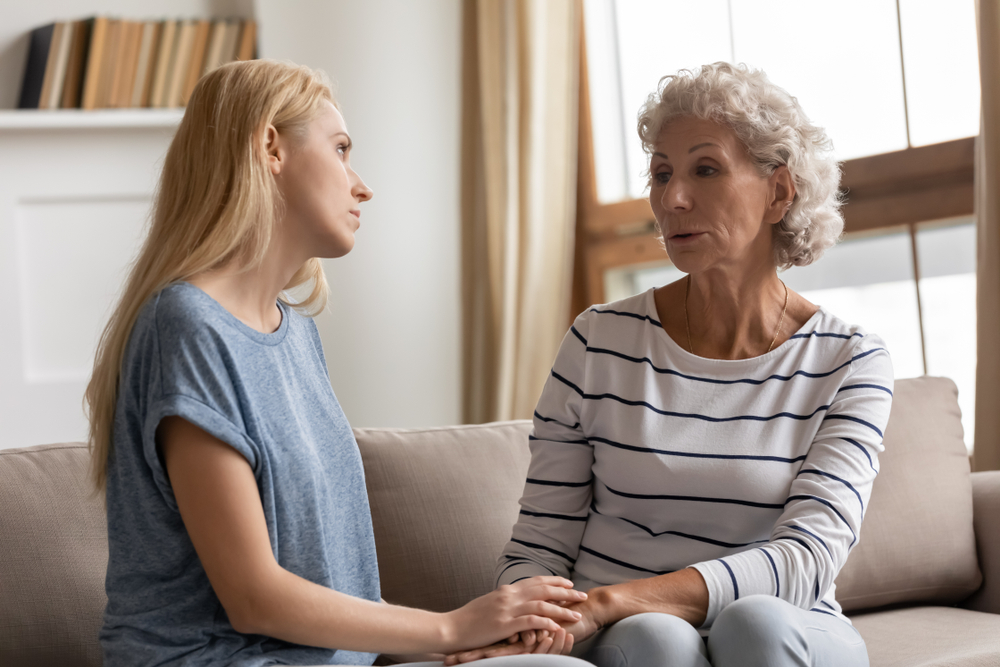 Compassionate grown up daughter listening to complaining old senior mother, sitting together on sofa. Loving adult kid holding hands of stressed middle aged mommy, showing support love care.
