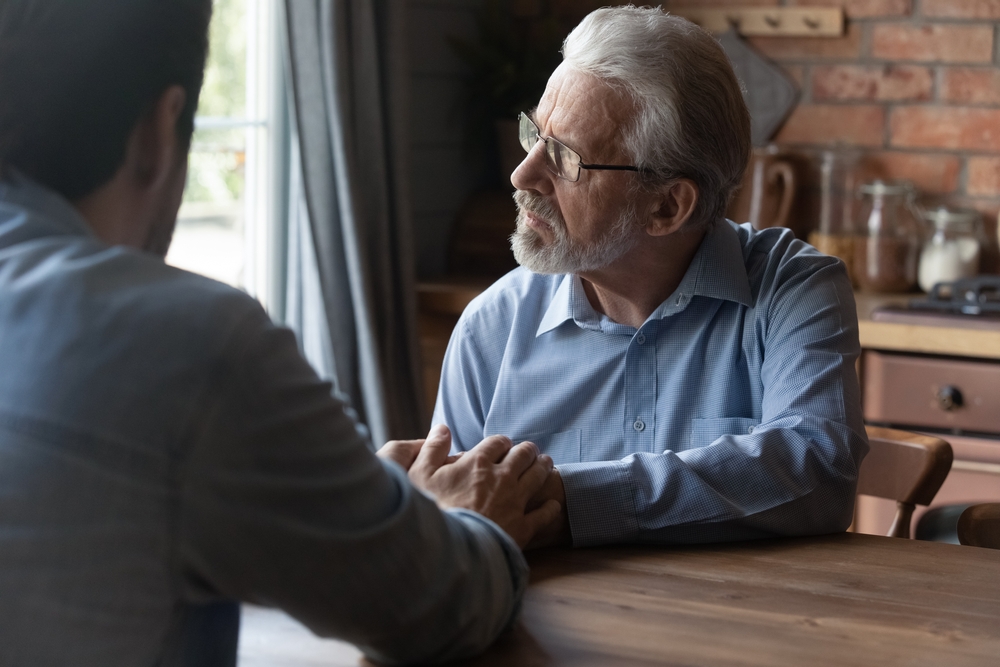 Helping hands of son. Attentive grownup kid sit at table close to sad senior hoary father touch his palms listen to problem. Young grandson give support to depressed old grandfather help overcome loss
v