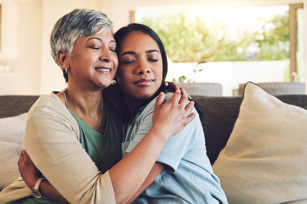 Family, senior woman hugging her daughter and love with people sitting on a sofa in the home living room during a visit. Smile, trust or comfort with an elderly female parent embracing an adult child
