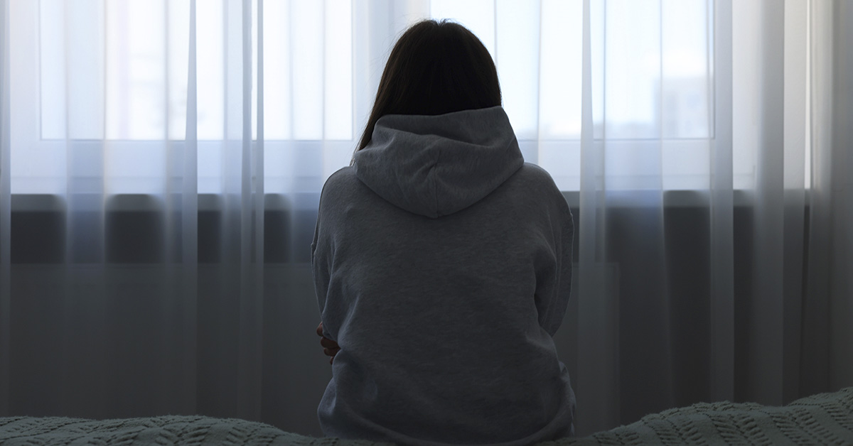 woman facing away sitting edge of bed looking out a window through sheet curtains