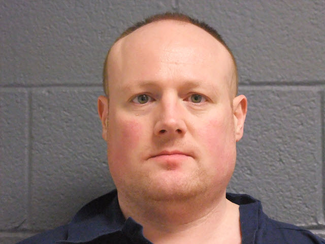 Booking photo of a white man with a shaved head wearing a blue prison uniform, photographed against a gray concrete block wall.