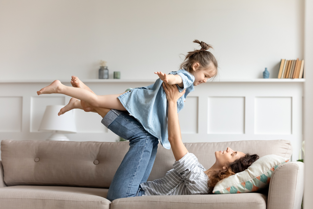 Active time with preschool child at home, easy comfort travel together with kid concept. Mother lying on couch lift up little daughter she raised hands imagines herself like plane enjoy flying in air