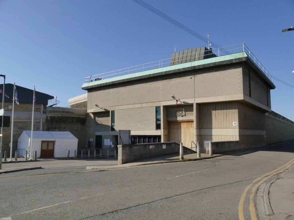 Exterior view of HM Prison Wakefield showing the back lane entrance. The building features beige brick walls, a fortified entrance with Royal coat of arms, security fencing on the roof, and a guardhouse at the front.