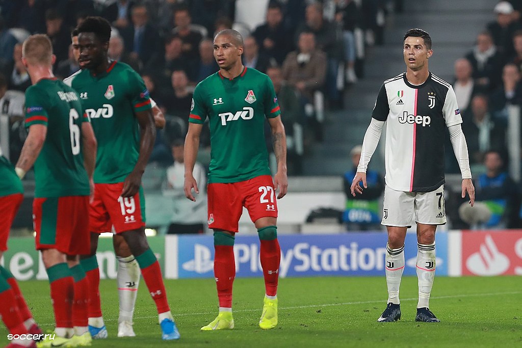 Cristiano Ronaldo in Juventus kit standing on pitch during match against Lokomotiv Moscow players in green and red uniforms.