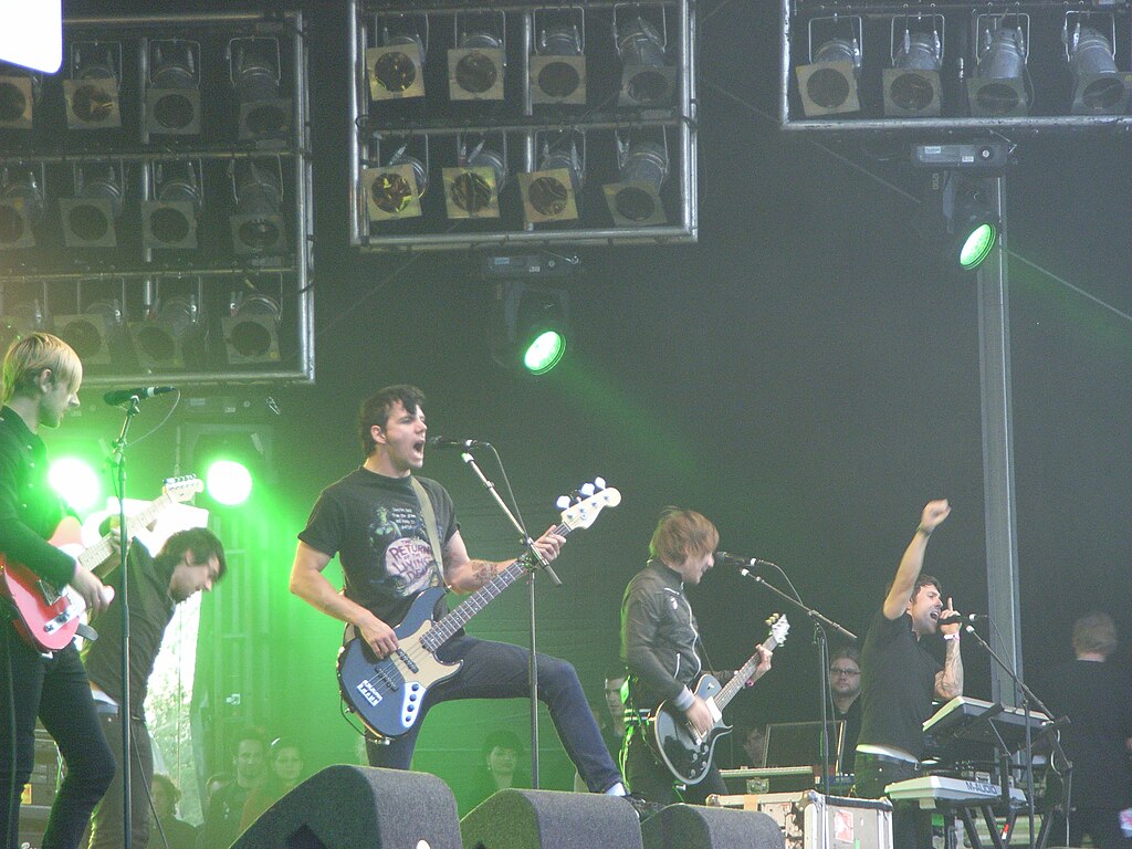 A rock band performing on an outdoor stage with green stage lighting. Five band members are visible, including guitarists, a bassist, and a vocalist with raised arm. Large speaker stacks are mounted above the stage.