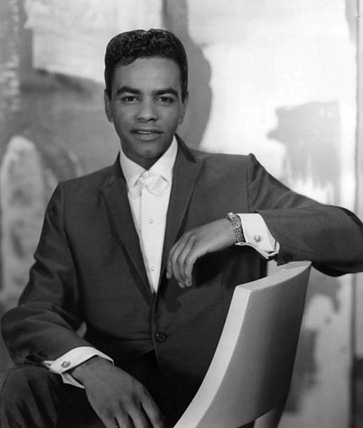 Black and white portrait of Johnny Mathis in 1960, a young man in a dark suit and white shirt, seated and looking at the camera.