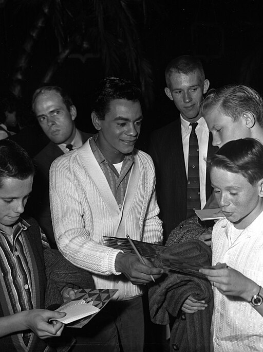Black and white photo of Johnny Mathis signing autographs while surrounded by several young fans reaching toward him with papers and albums.