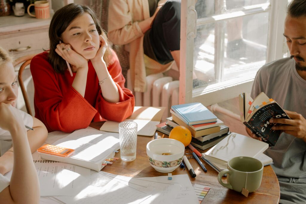 Woman in red sweater resting her head in her hands at a study table with others working nearby.