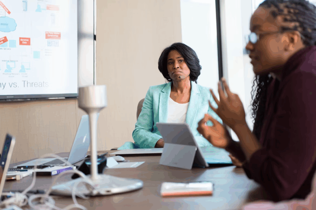People in a business meeting, one woman listening, appearing unimpressed, while another person gestures.