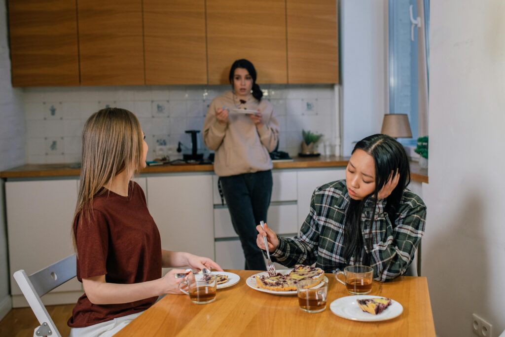 Three people in a kitchen, two seated at a table eating while one stands in the background.