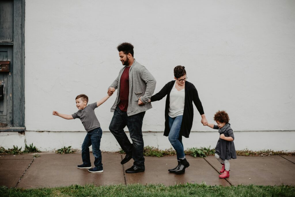 Two adults and two young children hold hands as they walk together down a sidewalk in front of a white building, all smiling and moving playfully.
