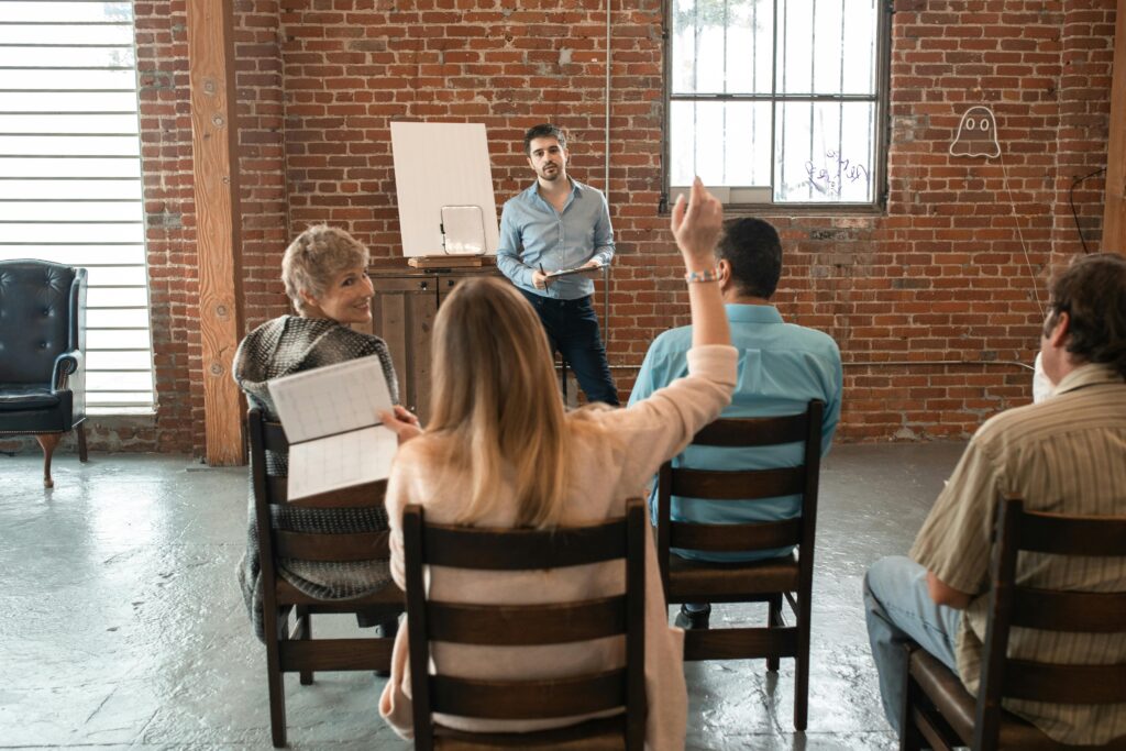 Man presenting while woman raises hand to ask question. Another woman seated left looks back at her, and a third person faces forward.