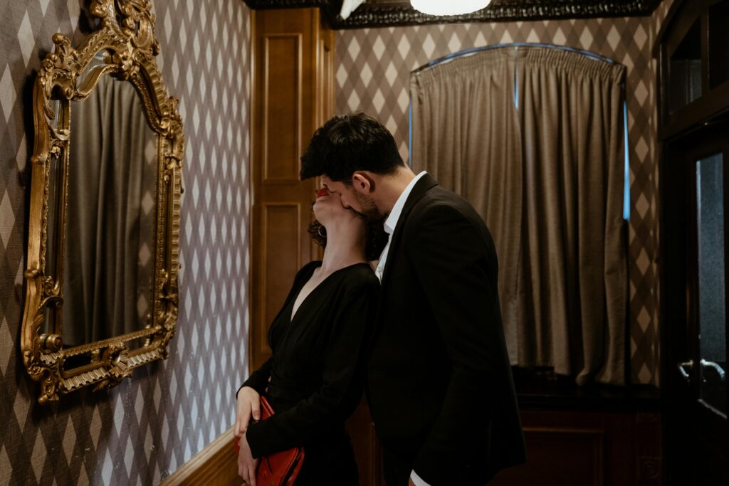 Man in Black Suit Kissing Woman in Black Dress Kissing on the Neck in Front of Mirror
