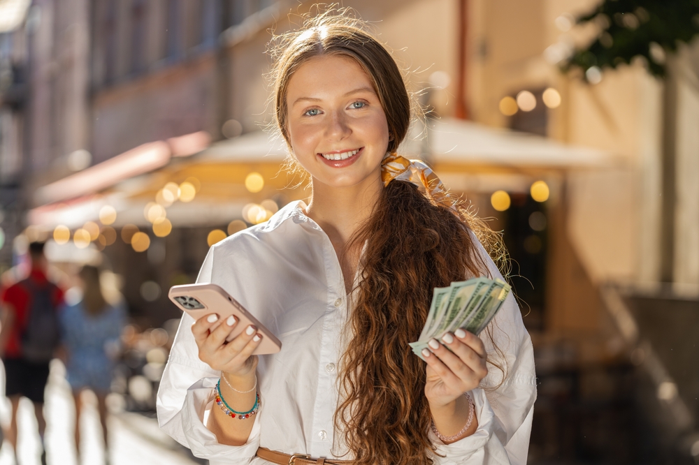 Happy rich teenager girl counting holding money dollar cash, use smartphone calculator app, plans to order gifts and food delivery online booking hotel room. Redhead young woman tourist on city street