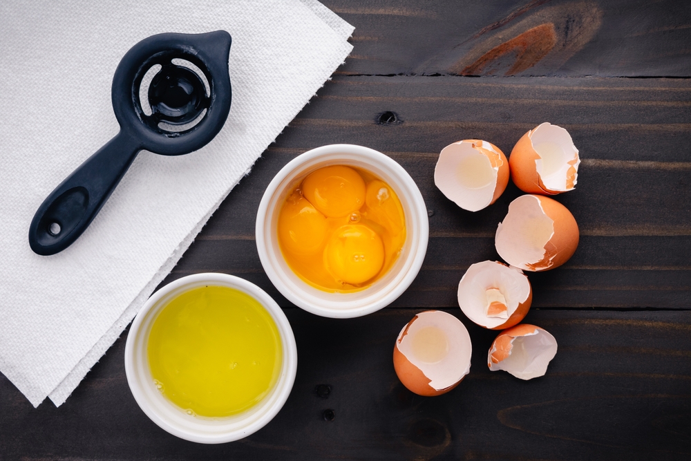 Four Separated Eggs in Small White Ramekins: Egg yolks and egg whites shown with an egg separator and egg shells on a wooden table