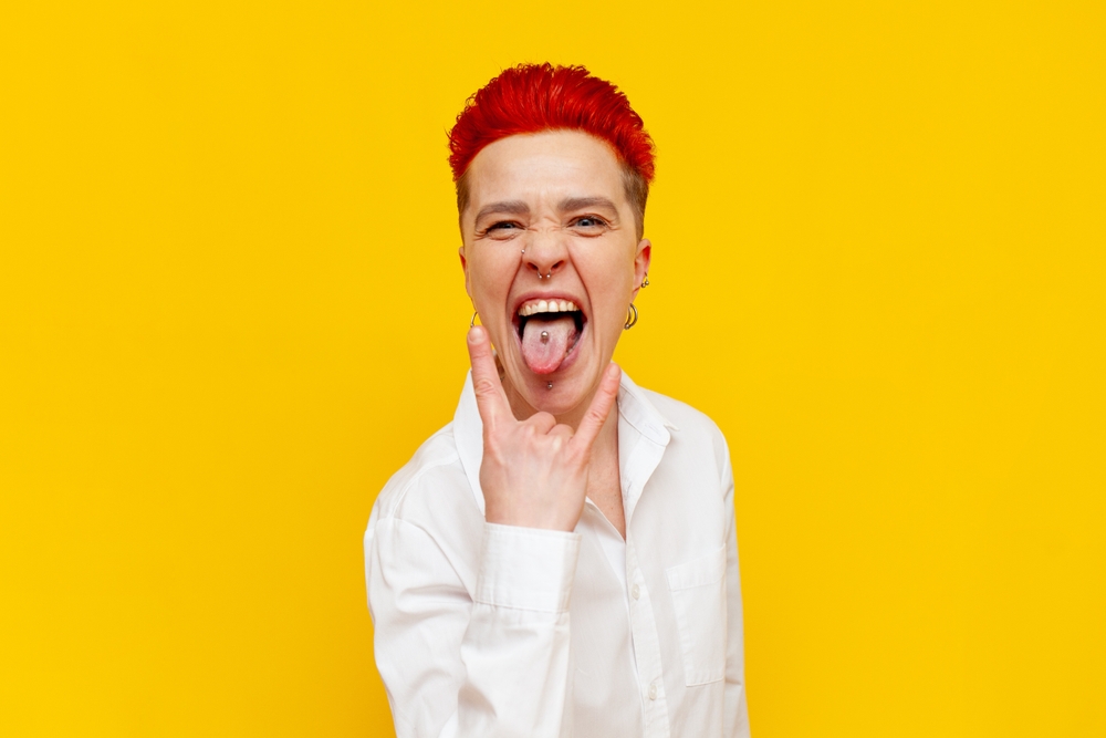 young short-haired red-haired punk girl shows rock gesture and screams on yellow isolated background, crazy hipster woman with piercing shows tongue with piercing