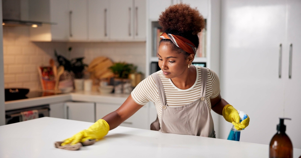 Woman, cloth and wipe on kitchen counter for hygiene with spray, gloves or chemical for shine in home. African person, cleaning and bottle for dust, dirt or bacteria with sanitation liquid in house