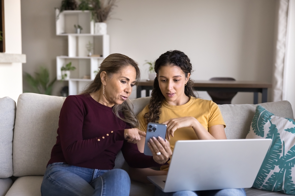 Sharing information in digital age. Friendly family mature mum teen daughter rest on sofa using diverse gadgets exchange social media content between phone notebook discuss travel plans shopping ideas