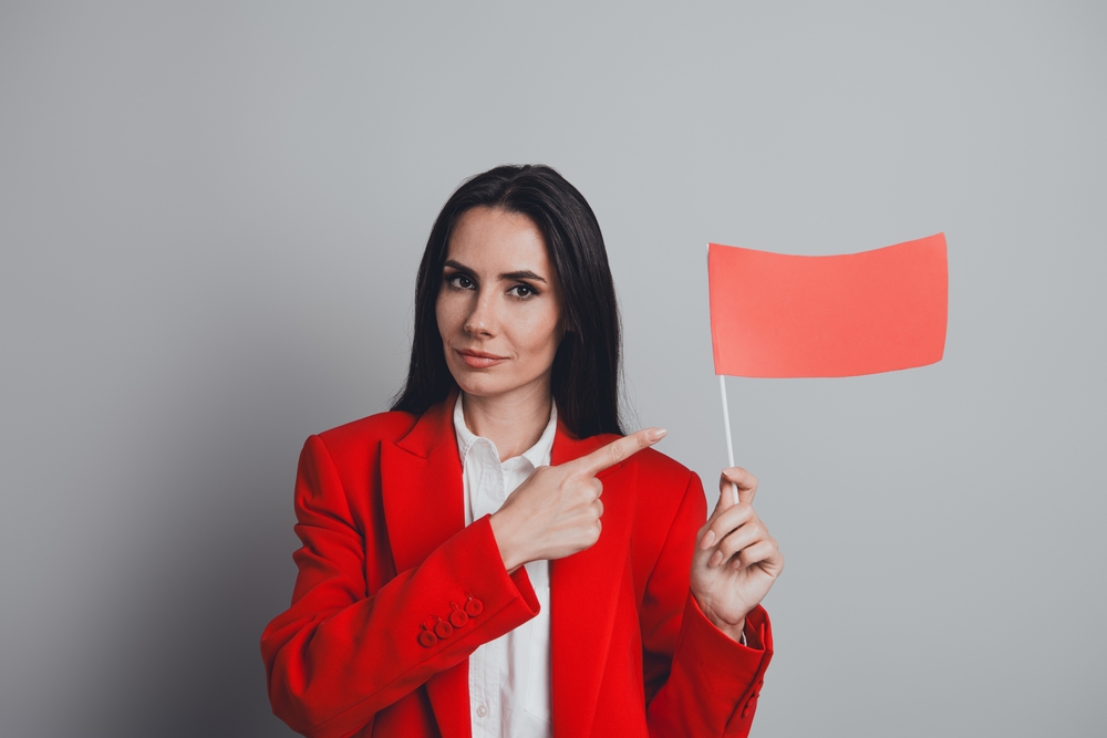 Successful businesswoman holding a red flag on a grey background showing a sign of alert, dressed in a red suit