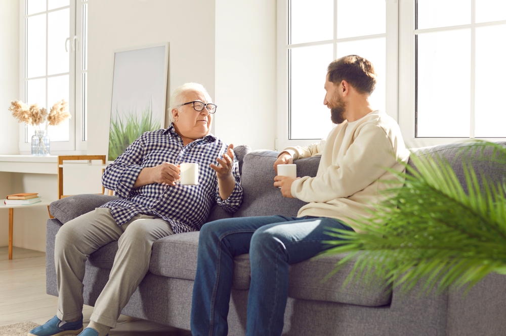 Adult son visiting his old father. Young man and his senior retired father sitting on the couch at home, drinking coffee, and talking. Family, communication concept