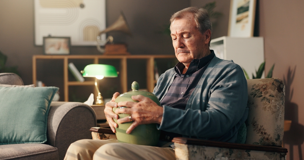 Senior, sad man and home with urn or memory for grief, loss or mourning in thought on chair. Elderly, male person or widower thinking with vase of ashes for nostalgia, missing piece or lonely house