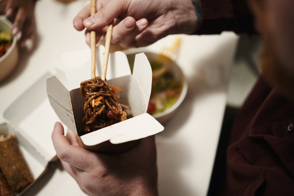 Hands of young man eating noodles with chopsticks from takeout box, holding container in one hand while lifting food, other people and bowls of food visible in background