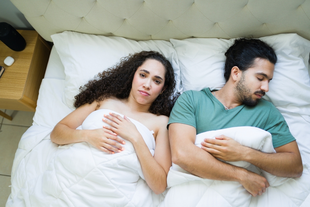Woman lying awake in bed, feeling restless and troubled while her partner sleeps peacefully beside her, highlighting relationship struggles