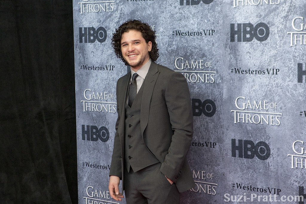 Kit Harington smiles on a red carpet, his curly dark hair framing his face. He wears a charcoal gray three-piece suit with a matching tie and light gray dress shirt, one hand casually tucked in his pocket. The step-and-repeat backdrop behind him features repeating HBO logos, Game of Thrones branding, and the hashtag #WesterosVIP in gray tones.