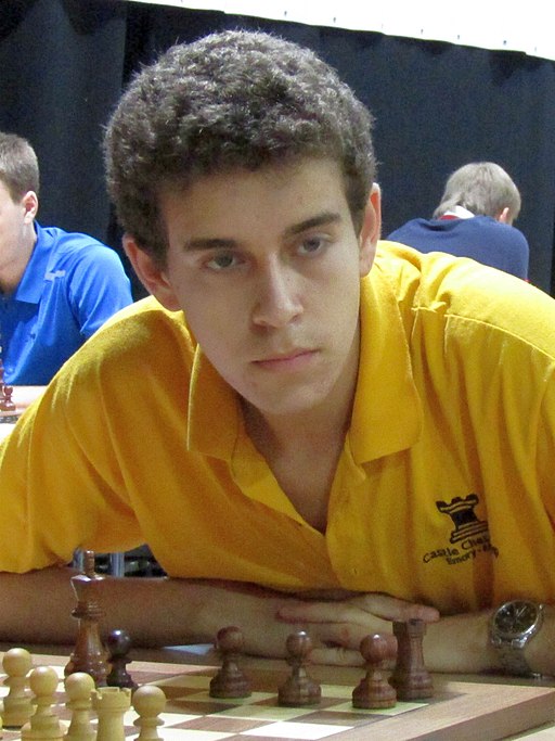 Daniel Naroditsky in a yellow polo shirt sits at a chess board with white pieces, looking directly at the camera during the 2016 RTU Open in Riga.