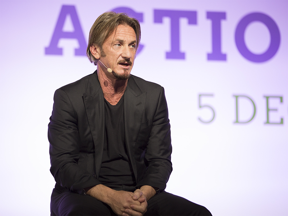 Sean Penn sits on stage during a panel discussion, wearing a black blazer over a black v-neck t-shirt. He has a small lavalier microphone clipped near his ear and his hands are clasped in his lap. His graying brown hair is pushed back from his face and he appears mid-sentence, looking slightly off-camera. Behind him, a purple and green backdrop shows partial text reading "ACTIO" and "5 DE."