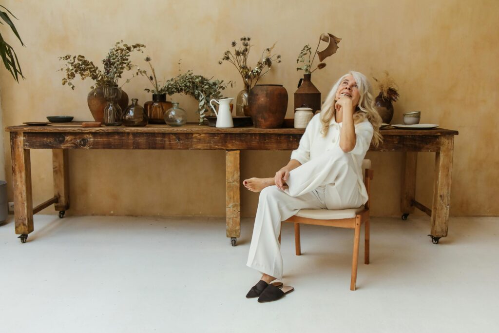 A woman with gray/silver hair in comfortable white clothing sitting on a wooden chair in a serene room with plants, pottery, and rustic wooden furniture. She looks peaceful and content.