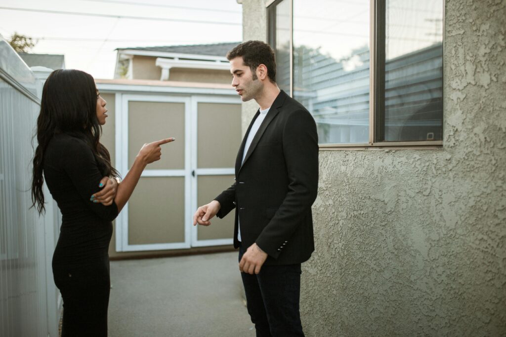 Woman in black demonstrating confident communication, pointing at man in suit who looks down, standing outside near a building.