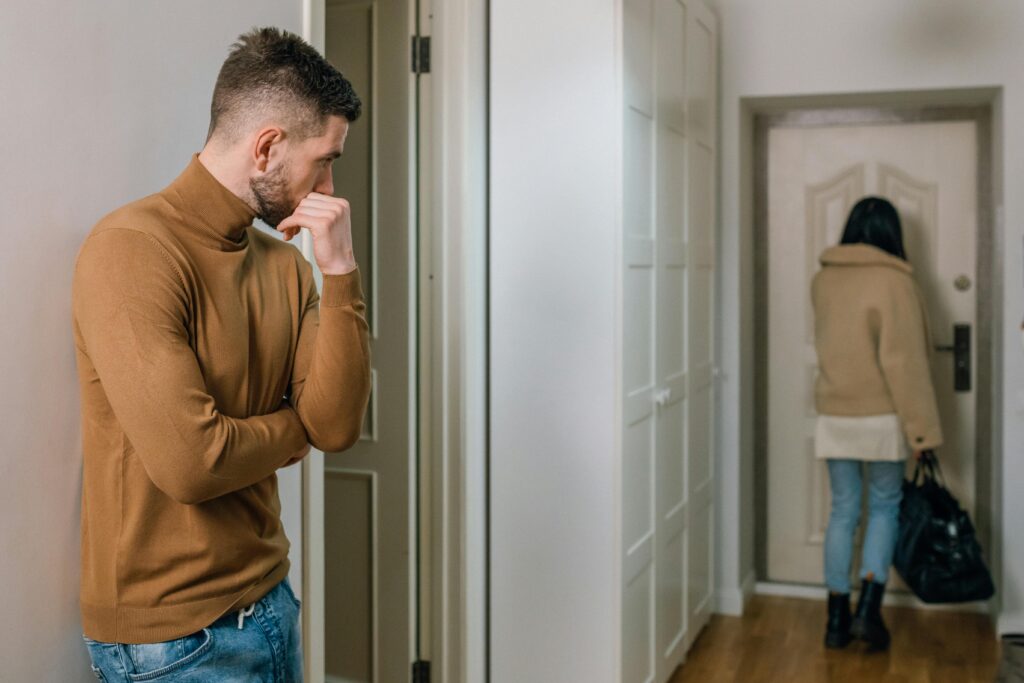 Man stands in hallway looking down while woman walks away toward door carrying a bag.