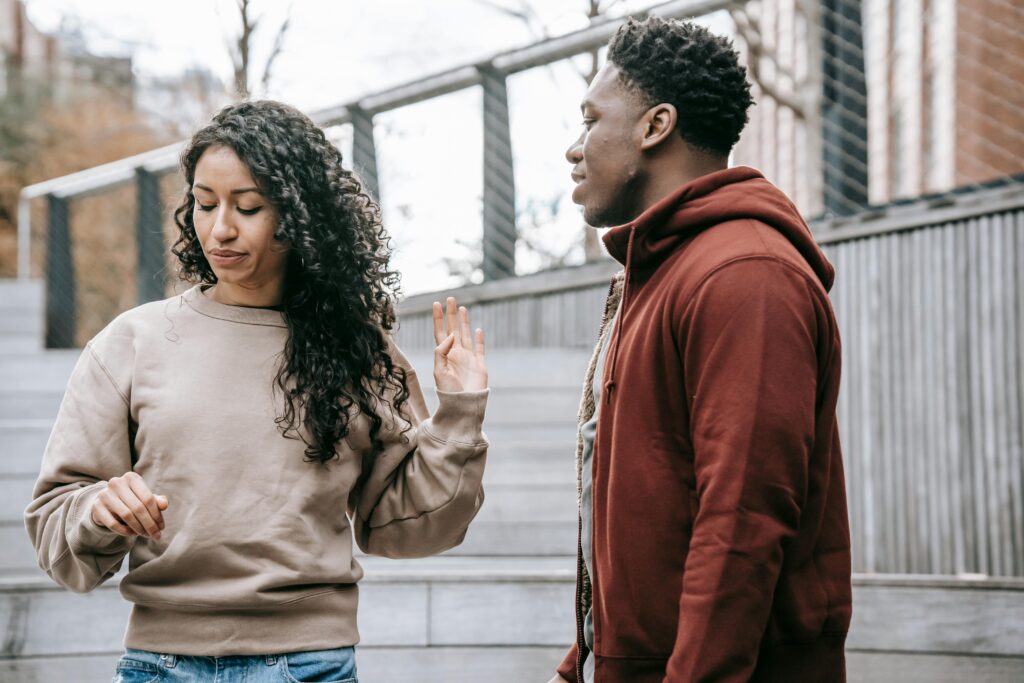 Woman with curly hair holds palm up in stop gesture toward man in red hoodie, outdoors on steps.