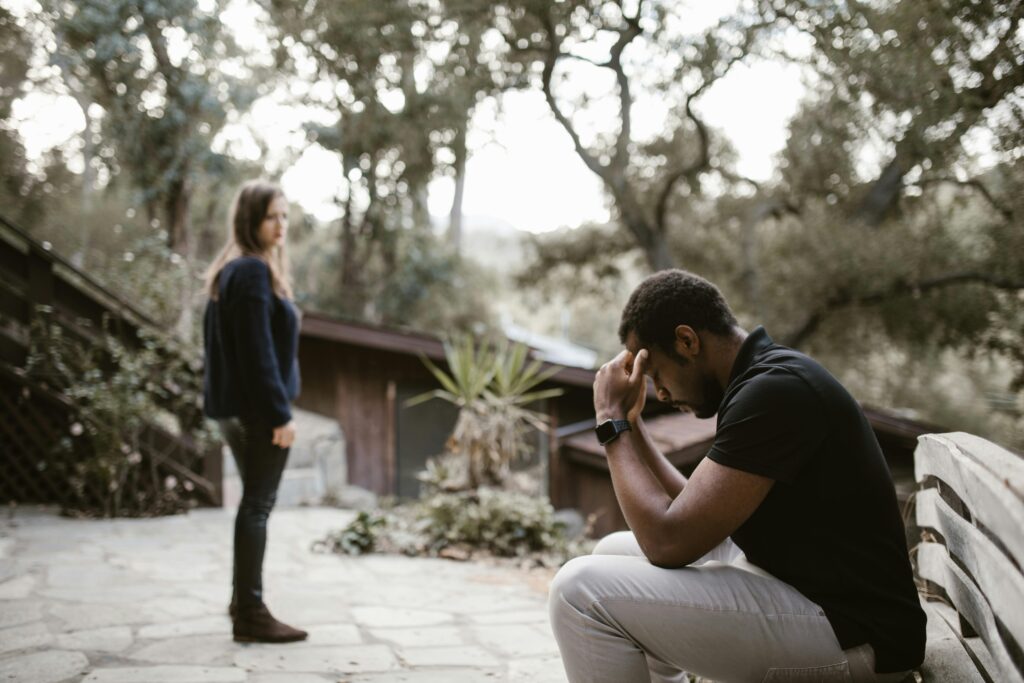 Man sits in foreground with head in hands while woman stands at a distance behind him looking back, they are outdoors trees.
