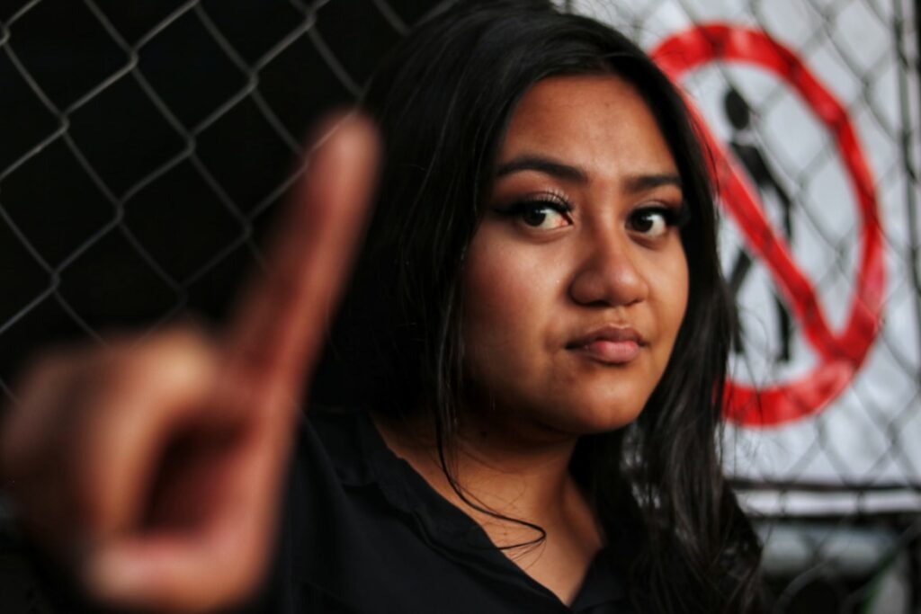 Young woman with a serious expression holding up one finger toward camera, a no walking sign visible behind her.