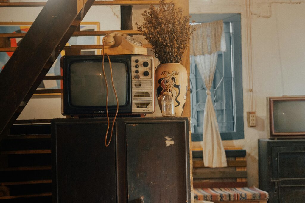 Old TV, rotary phone, and a vase with dried flowers sitting on a worn cabinet in a cluttered vintage room.