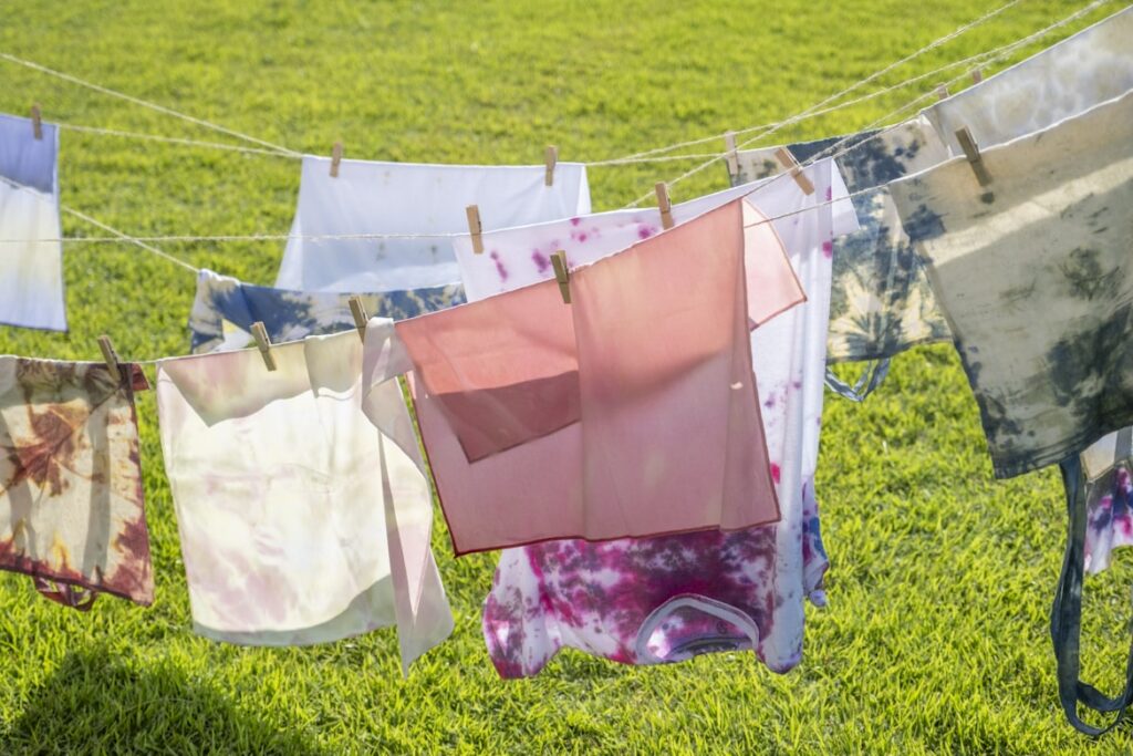 Colorful laundry hangs on a clothesline in bright sunshine, with tie-dyed fabrics, and pink cloth secured by wooden pegs over a grassy lawn.