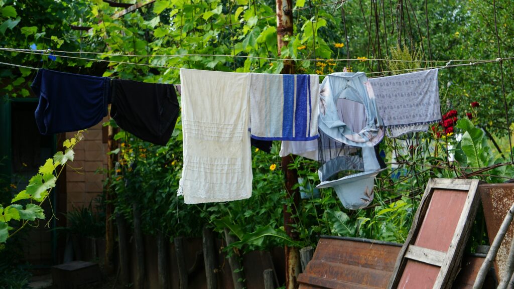 Laundry including a beekeeper's veil on a washing line in a leafy backyard garden, surrounded by towels in blues, blacks, and creams.