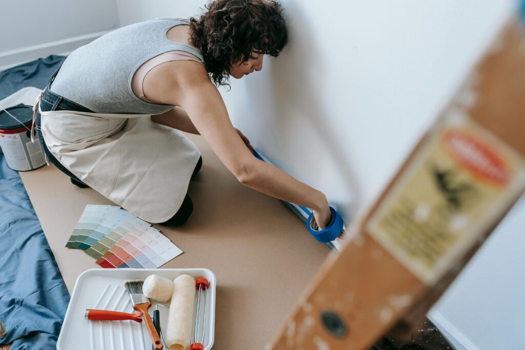 A woman kneeling on the floor, placing tape along a wall. She's wearing casual clothes with an apron, her hair pulled up. Behind her sits a bucket of paint, with color swatches nearby and a paint tray in the front.