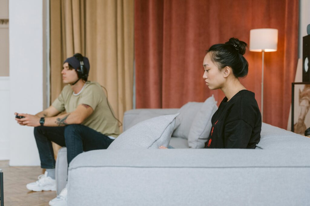 A couple sitting on a couch in a living room with warm orange curtains. The man wears headphones and a tan shirt, facing away and focused on something (likely gaming). The woman in a black top sits separately on the couch, looking distressed and lost in thought.