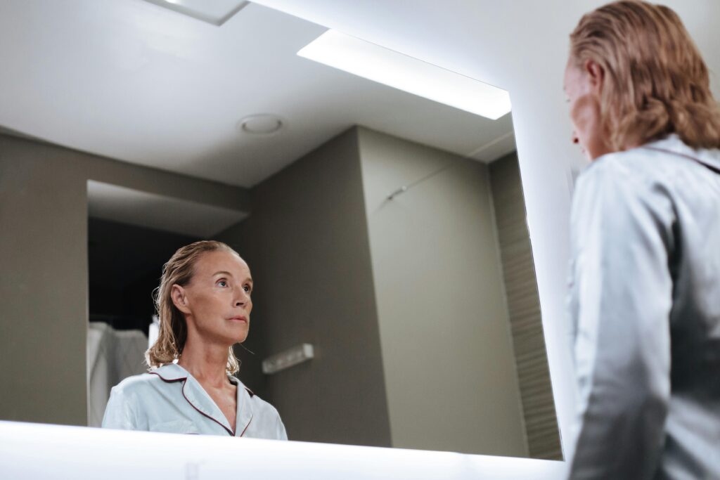 A woman in a light blue shirt looking at herself in a bathroom mirror. She has blonde hair pulled back and appears to be in a moment of self-reflection.