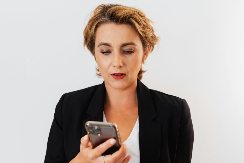 A woman with short blonde/light hair in a black blazer, looking down at her phone with an unimpressed/tired expression.
